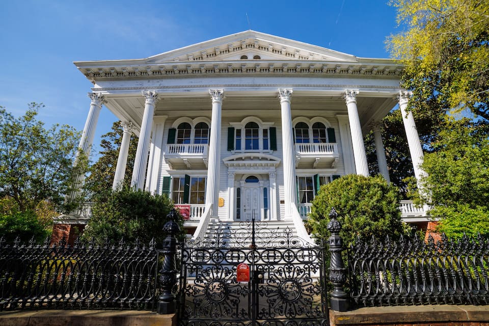 A white colonial-style building with columns and a balcony, surrounded by trees and a wrought iron fence.