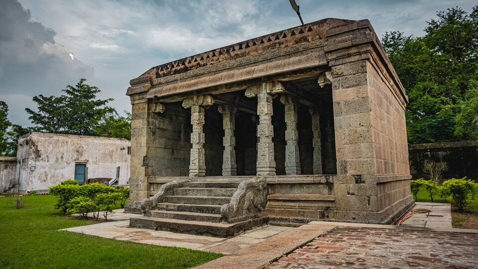 A stone temple with columns and steps, surrounded by greenery and a cloudy sky.