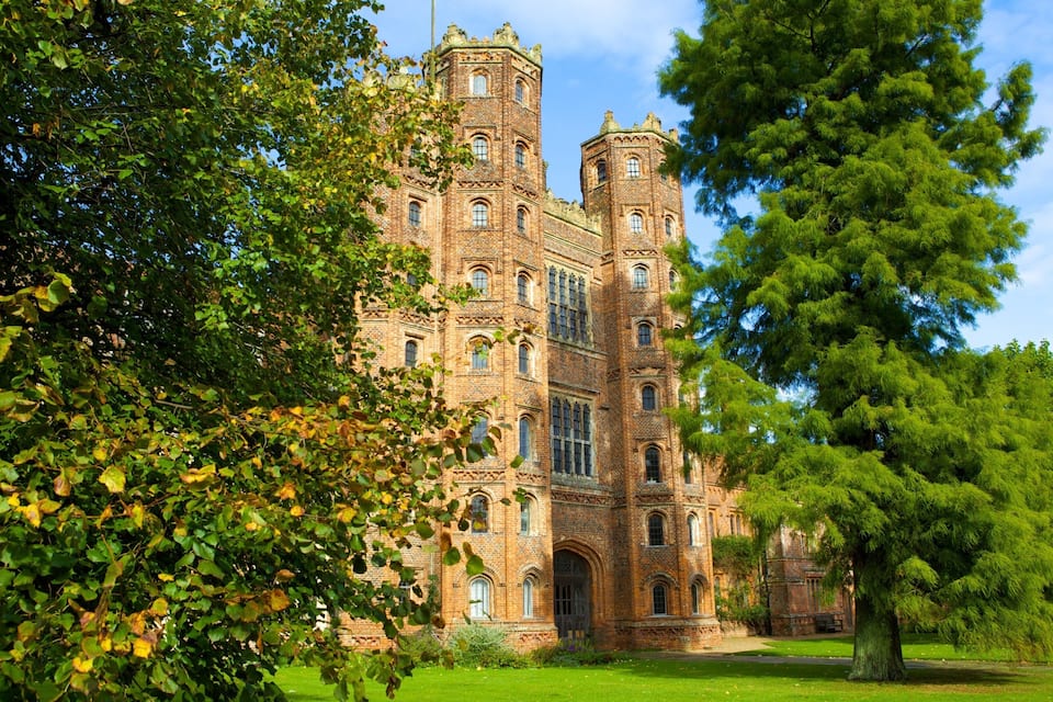 A large, historic brick building with multiple towers and arched windows, surrounded by lush green trees and a well-maintained lawn.