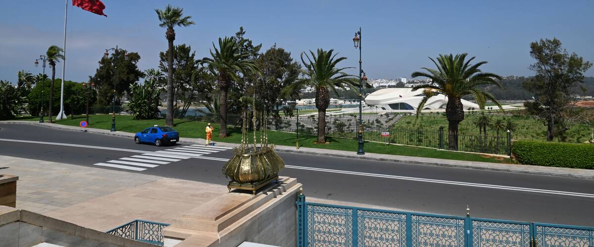 A road with palm trees, a blue car, a red flag, and a modern building in the background.