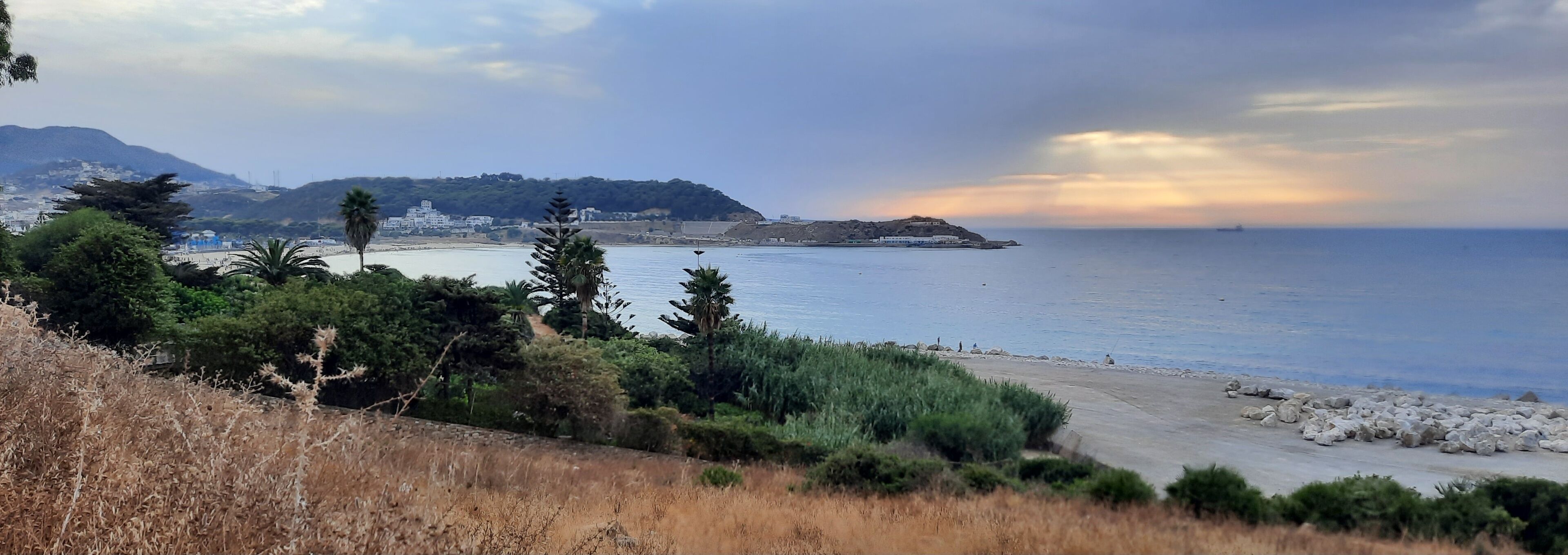 A coastal landscape with a beach, rocky shoreline, and a hill with buildings in the background.