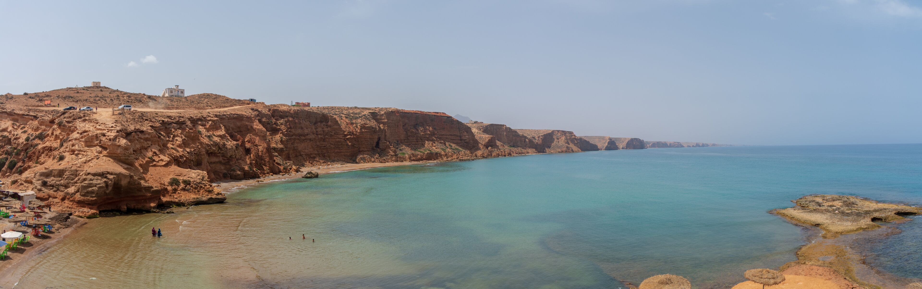 A coastal landscape with cliffs, clear turquoise waters, and a few buildings on the hillside.