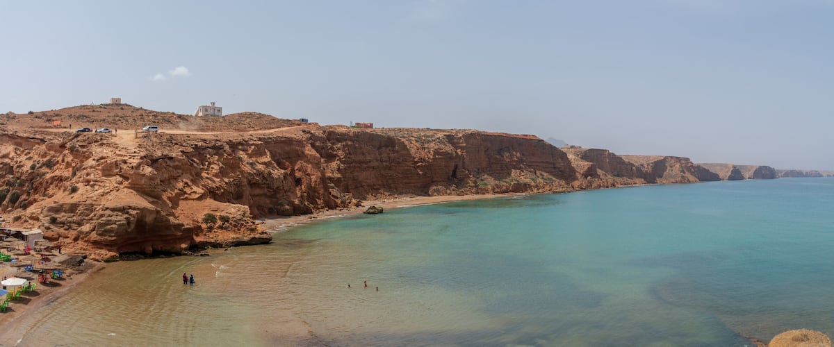 A coastal landscape with cliffs, clear turquoise waters, and a few buildings on the hillside.