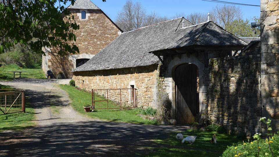 A stone building with a thatched roof, a gate, and a pathway leading to a larger stone structure with a tower.