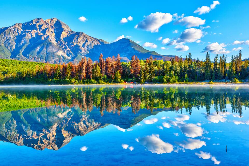Uma cordilheira, um lago com reflexo límpido e uma floresta de árvores em tons outonais.