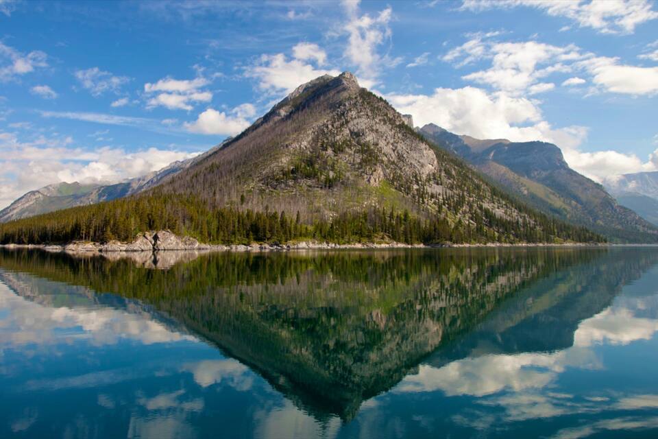 A mountain reflected in calm waters, surrounded by dense forests and under a partly cloudy sky.