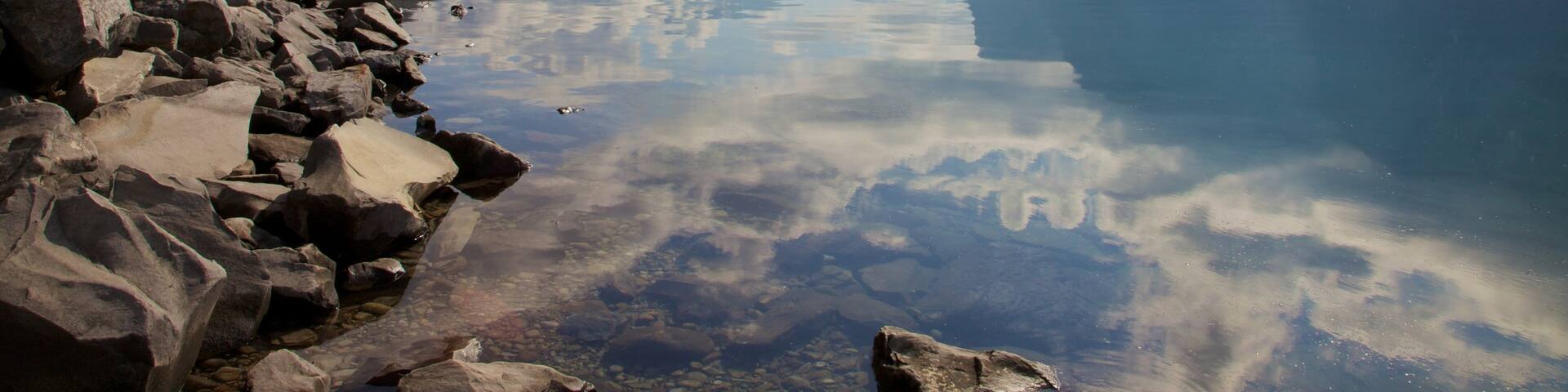 A calm lake with clear reflections, surrounded by mountains and rocky shores.