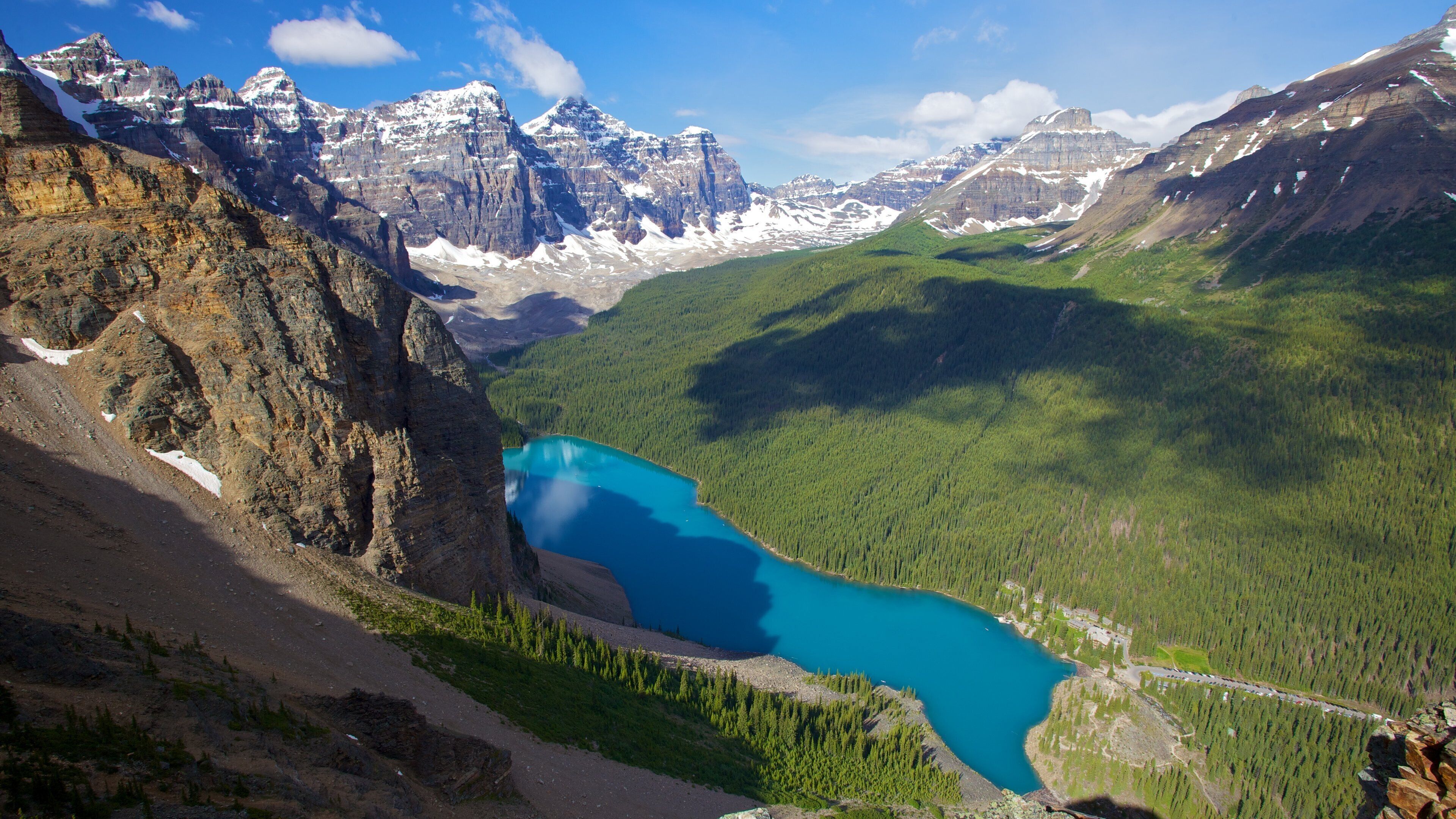 A mountain landscape with a turquoise lake surrounded by forest and snow-capped peaks.