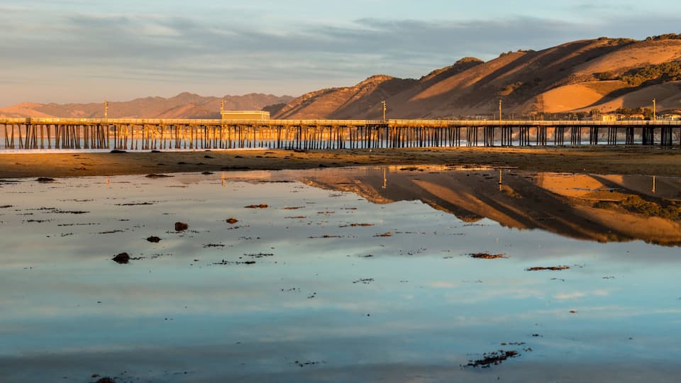 A pier extending into calm waters with a clear reflection, surrounded by hills and a clear sky.