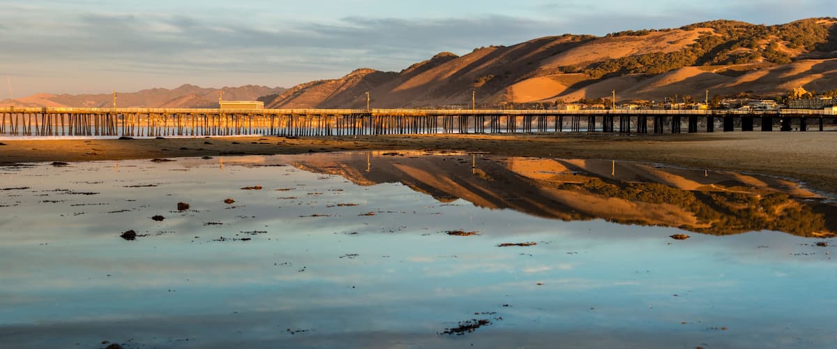A pier extending into calm waters with a clear reflection, surrounded by hills and a clear sky.