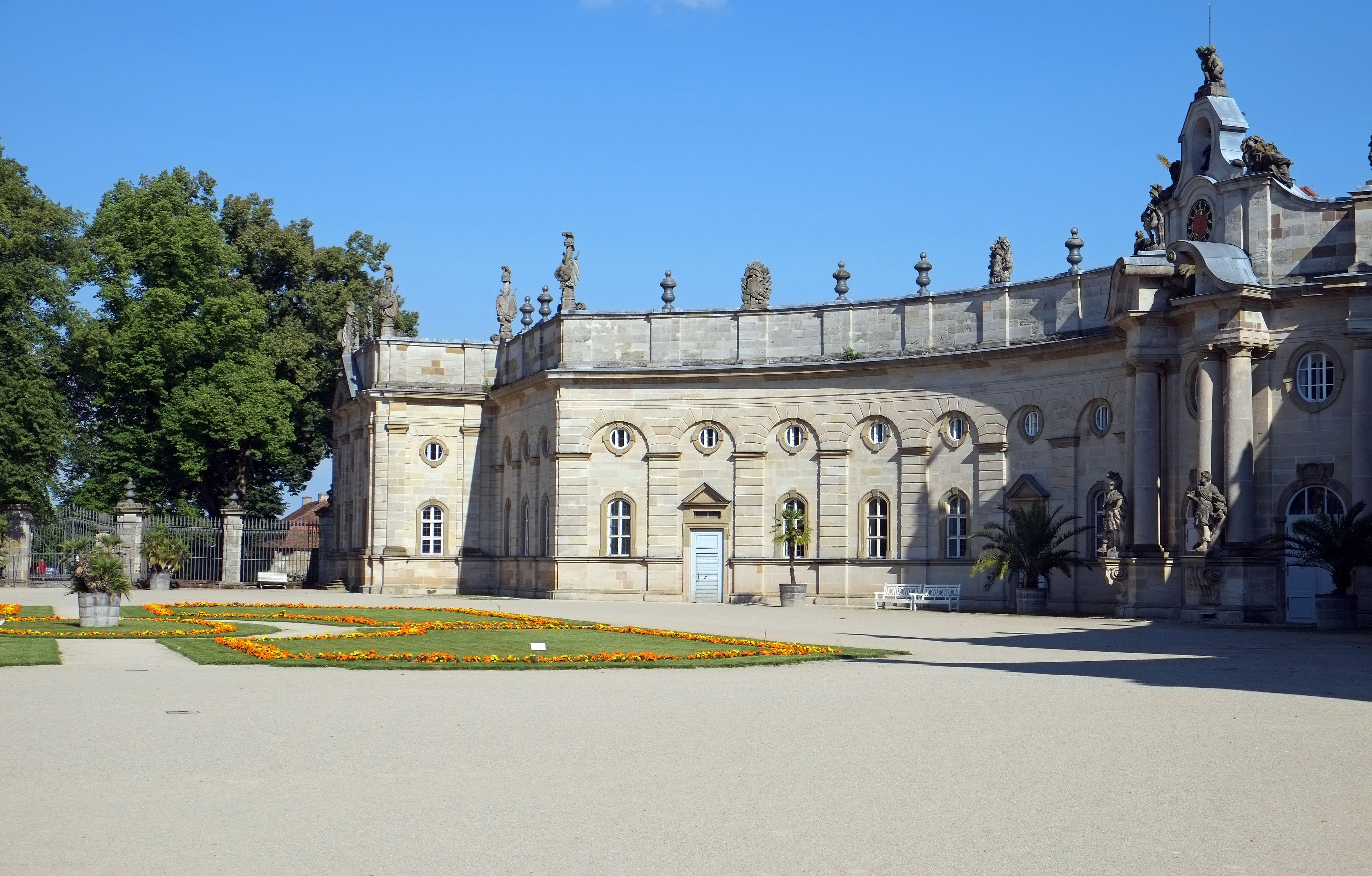 Un bâtiment historique à l’architecture classique, caractérisé par une grande porte centrale, des fenêtres en arc de cercle et des statues sur le toit.