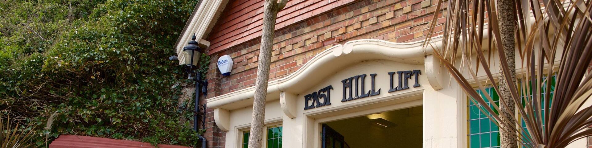 Un edificio con fachada de ladrillo, identificado con la inscripción “East Hill Lift”, una banca y follaje circundante.