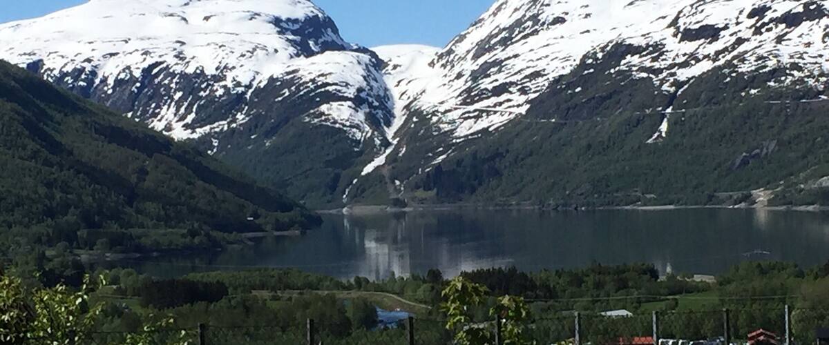 A lake surrounded by snow-capped mountains, a clear sky, and a fenced area with buildings.