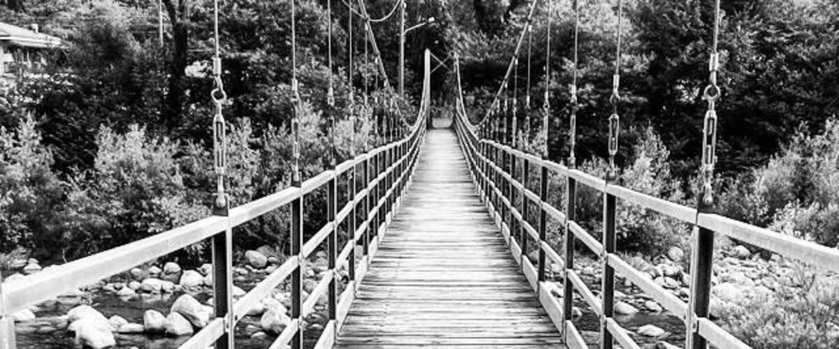 A wooden suspension bridge over a rocky stream surrounded by trees.
