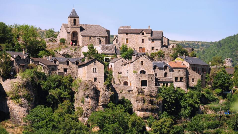 A historic stone village with a prominent church on a hillside, surrounded by lush greenery.