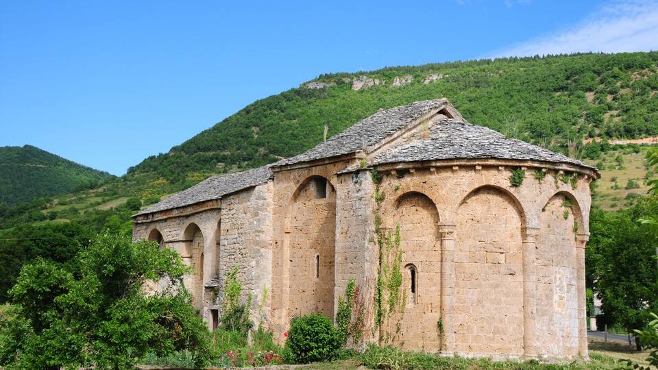 A stone building with arched windows and a tiled roof, surrounded by greenery and hills.