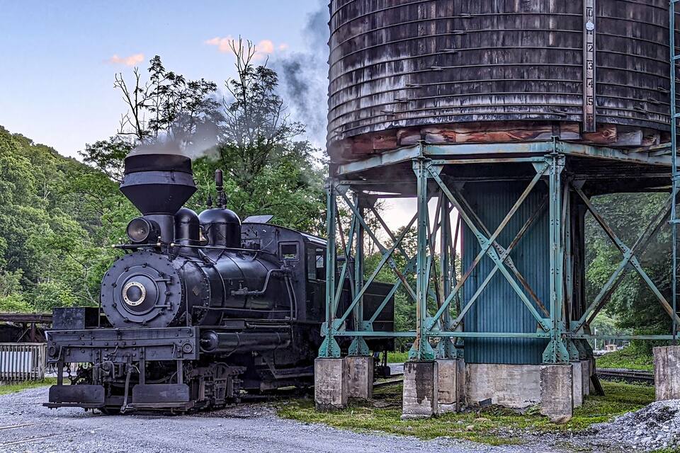 Een stoomlocomotief met een grote, cilindrische ketel die stoom uitstoot, naast een hoge, cilindrische watertoren die door een metalen constructie wordt ondersteund.