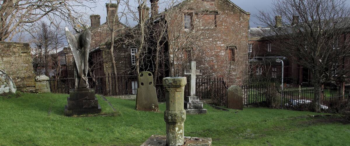 A cemetery with a stone cross, gravestones, and a brick building in the background.