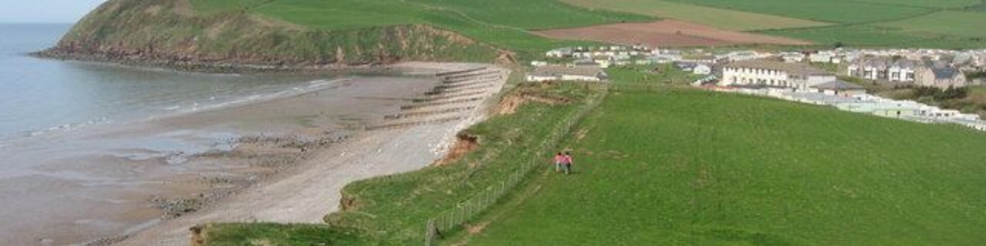 Un paisaje costero con una playa arenosa, campos cubiertos de hierba y un pequeño asentamiento con edificios blancos.