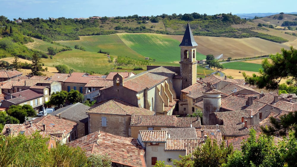 Un village aux bâtiments coiffés de toits en tuiles de terre cuite, une église dotée d’un clocher et, en arrière-plan, de douces collines verdoyantes.