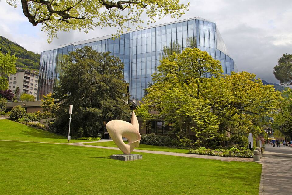A modern glass building, a sculpture, and a park with trees and a pathway.