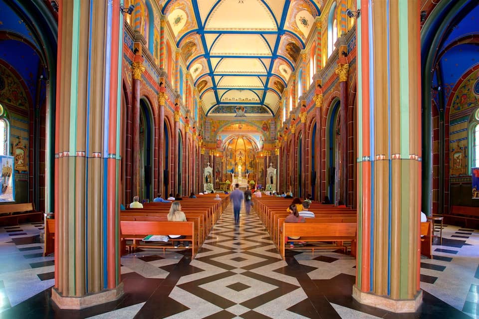 Interior de igreja com vitrais coloridos, um corredor central e bancos de madeira.