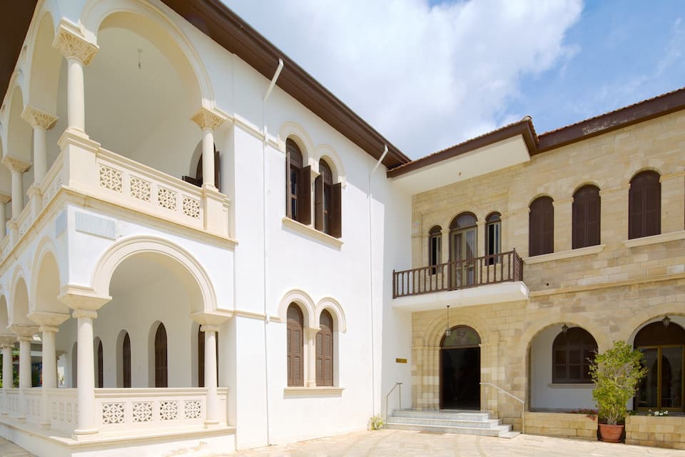 A white building with arches and a balcony, adjacent to a stone building with windows and a door.