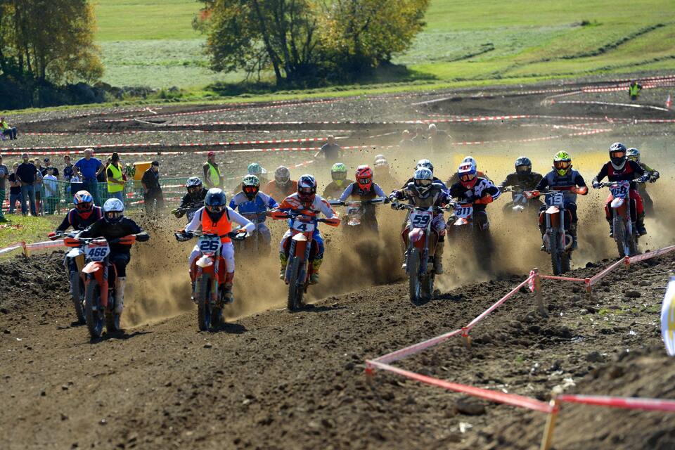 A group of motocross riders on a dirt track with spectators in the background.