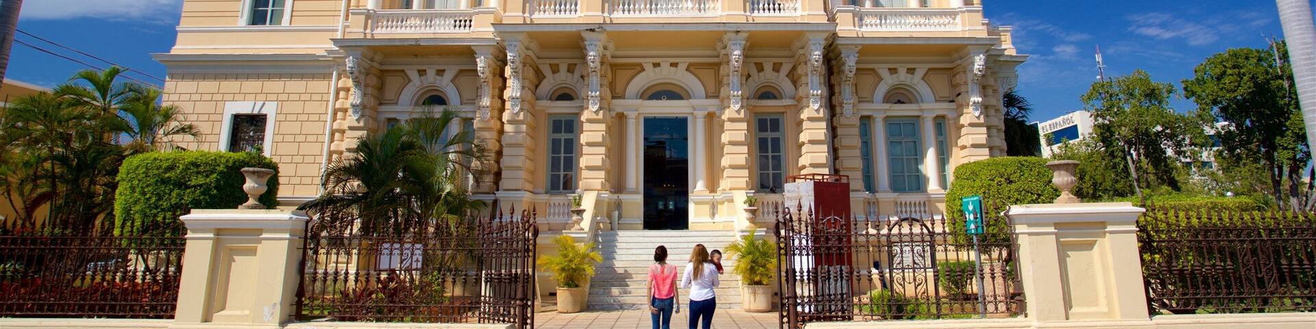 A grand, classical building with ornate columns and a symmetrical facade, surrounded by lush greenery and palm trees.