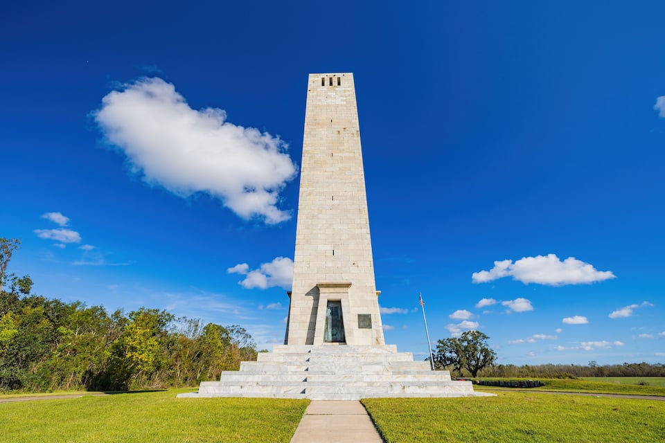 A tall, white monument with a flagpole, surrounded by green grass and trees under a clear blue sky.
