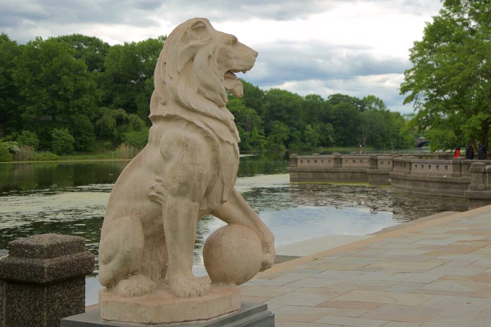 A stone lion statue on a paved walkway by a lake.