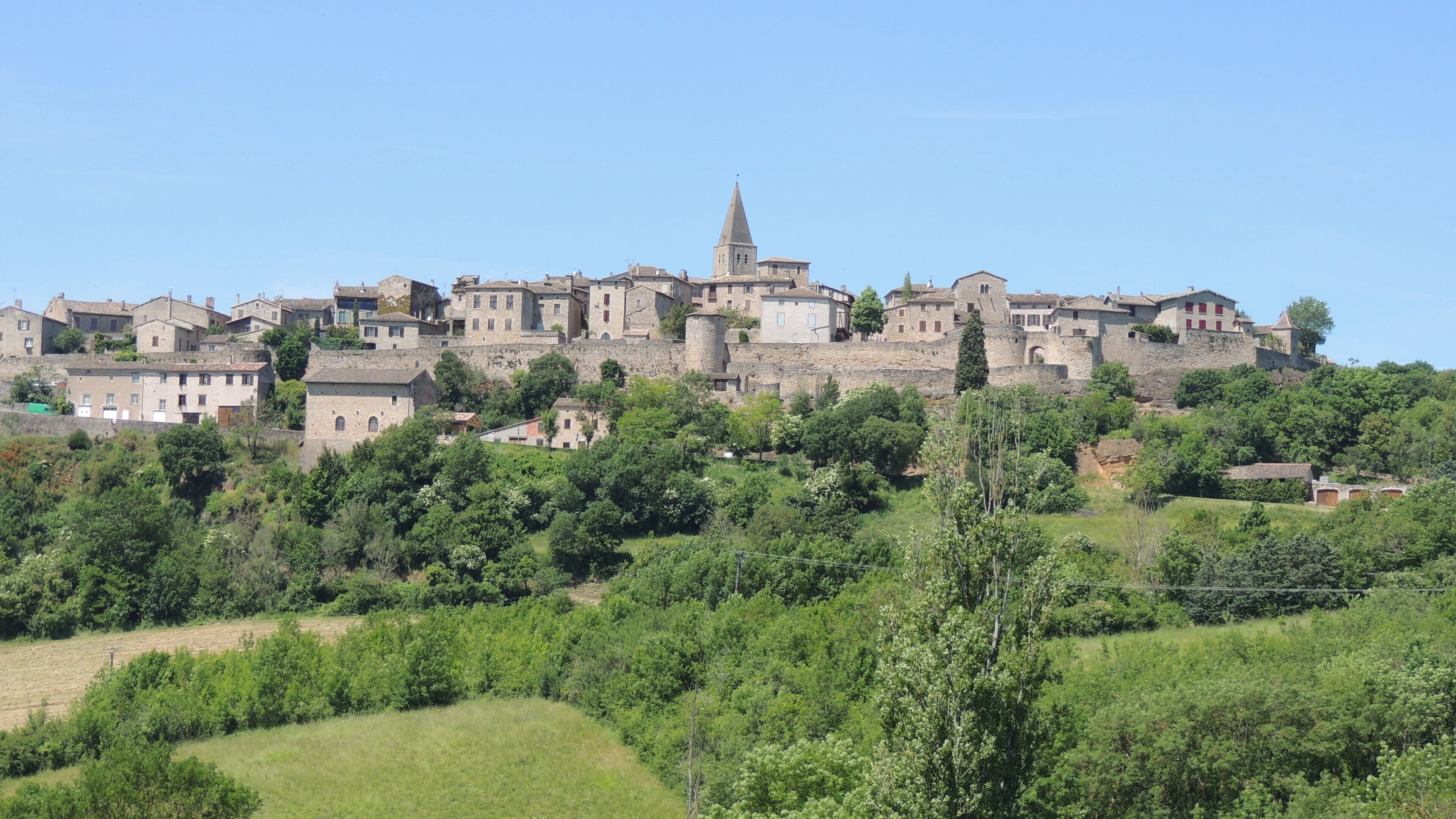 A historic town with medieval architecture, including a prominent tower and fortified walls, set against a backdrop of lush greenery.