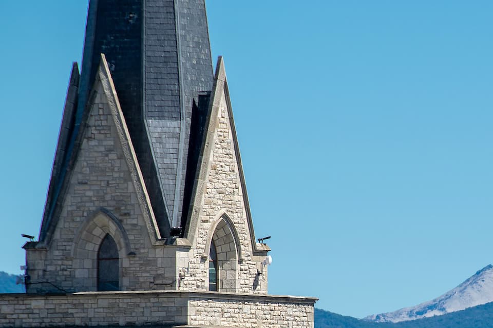A tall stone church with a pointed spire, set against a backdrop of snow-capped mountains and a clear blue sky.