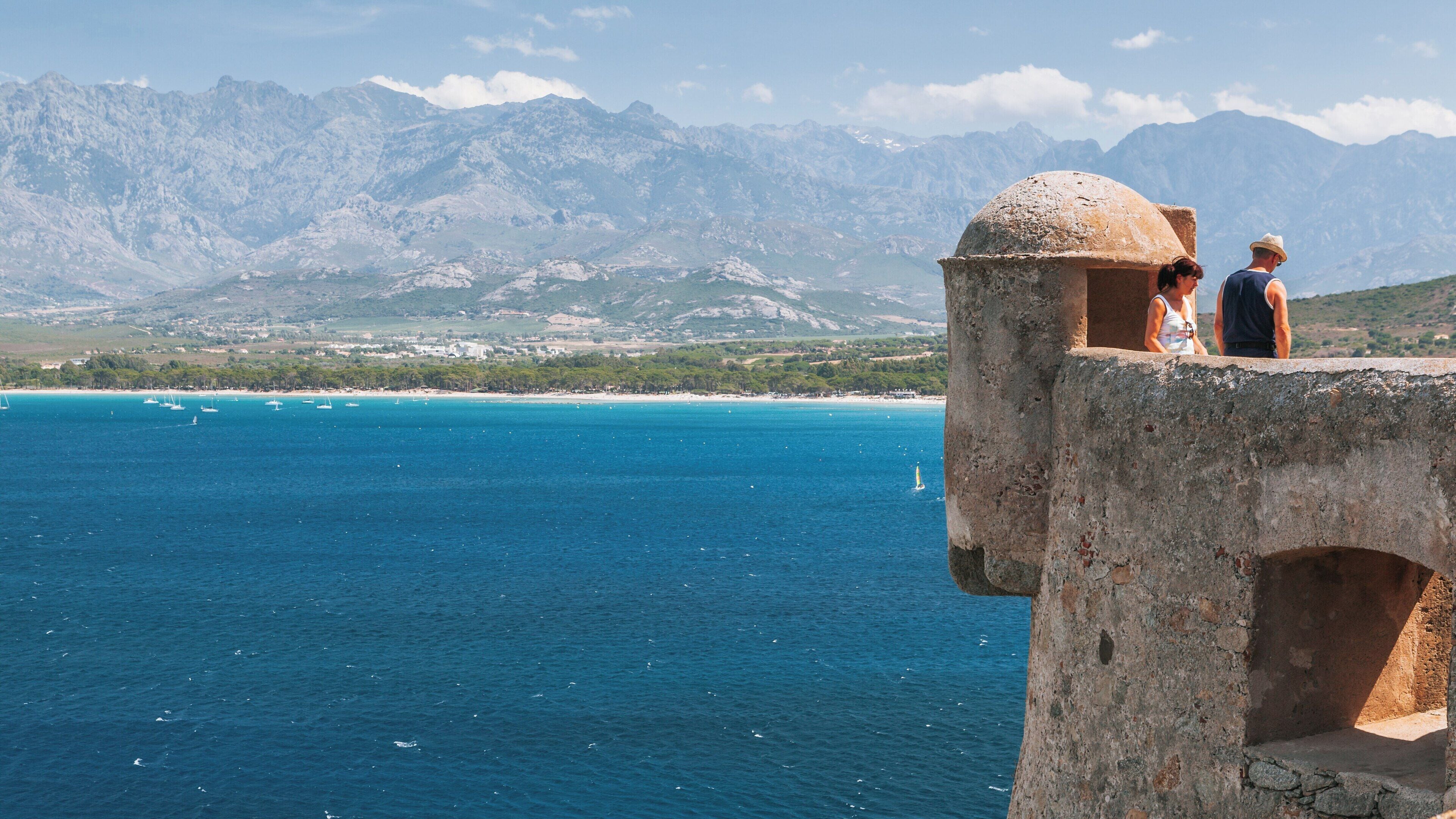 Visitors admire the view from Chapelle de Notre Dame de la Serra in Calvi, Corsica, with stunning backgrounds of mountains and the Mediterranean Sea