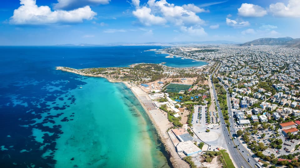 An aerial view of a coastal city with a clear blue sea, a sandy beach, and dense urban development.