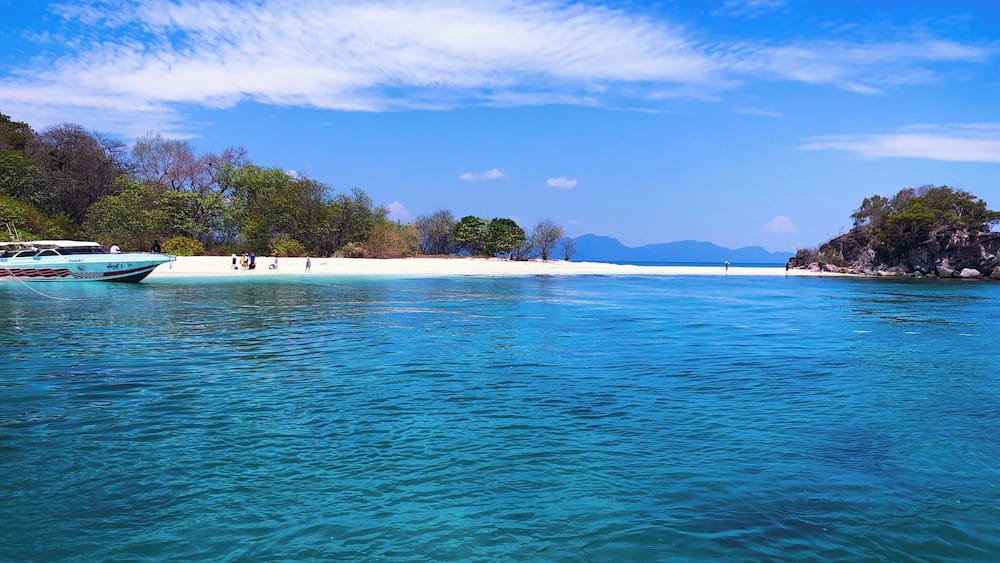 A clear turquoise sea with a boat named "FARIS 2" anchored near a sandy beach with people and trees.