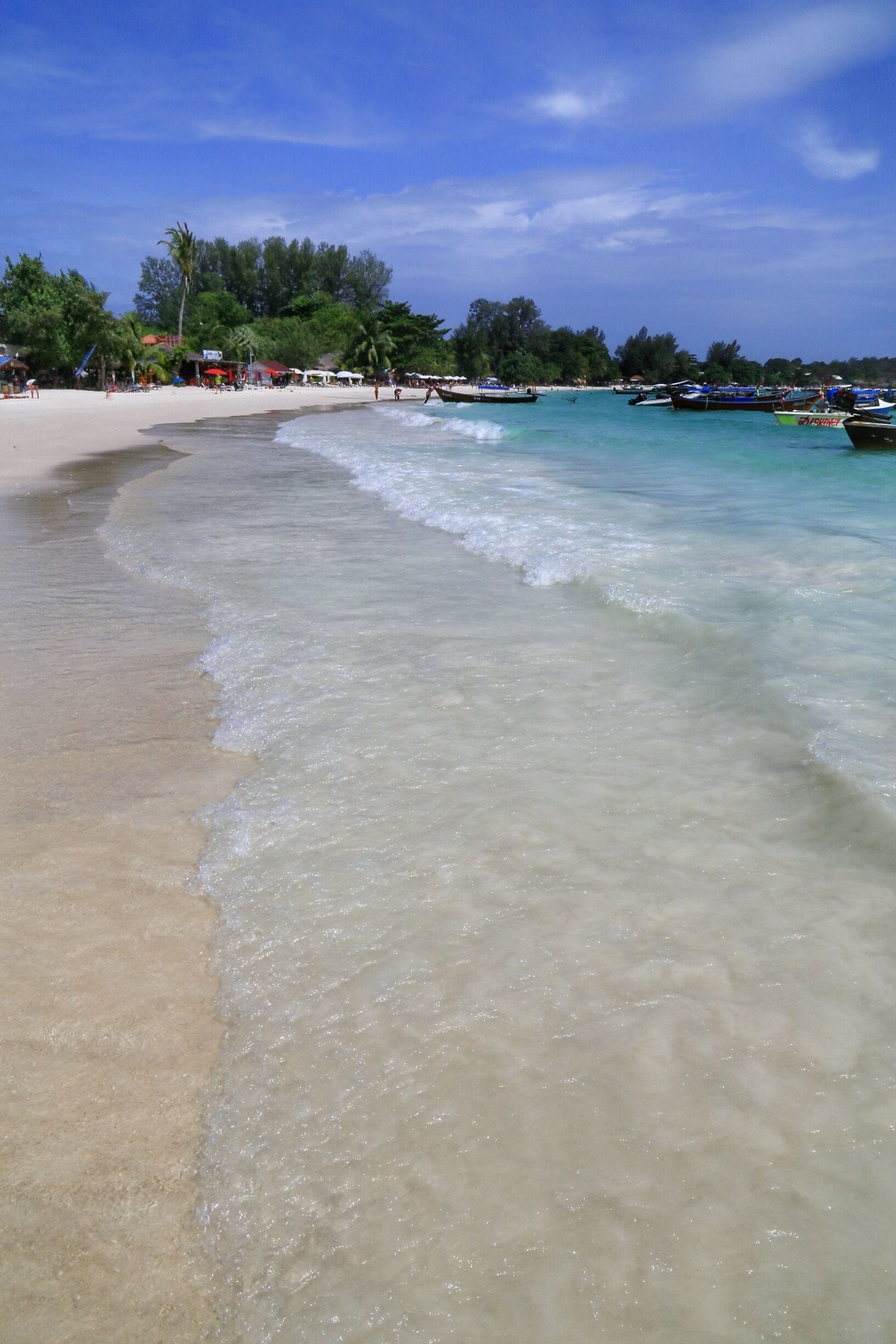 A beach with clear turquoise water, a sandy shore, and several boats anchored in the distance.