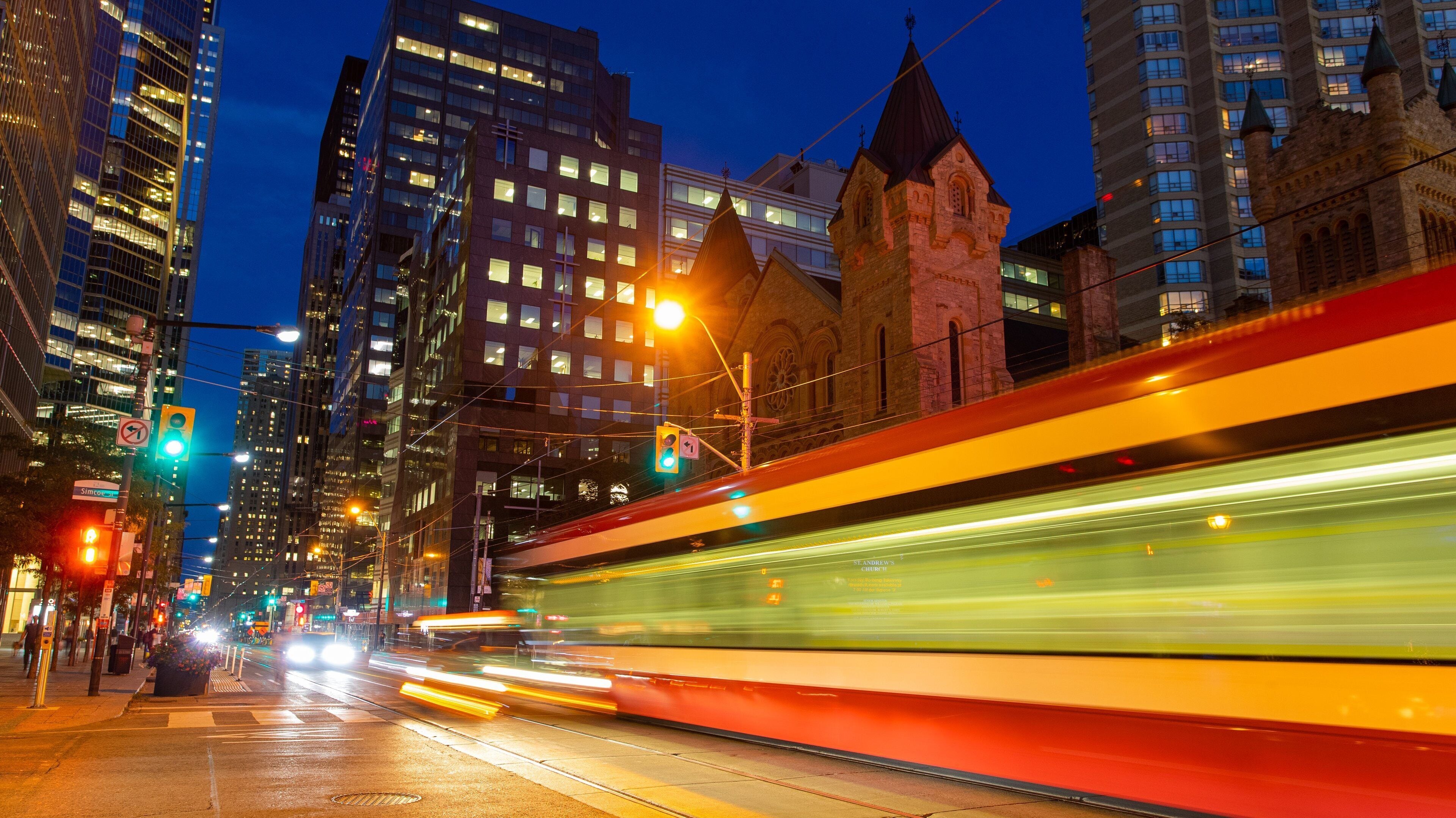A nighttime city street with a moving train, illuminated buildings, and a historic church tower.