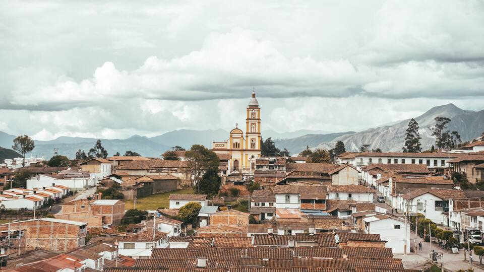 A town with terracotta-roofed buildings, a prominent church with a bell tower, and mountains in the background.