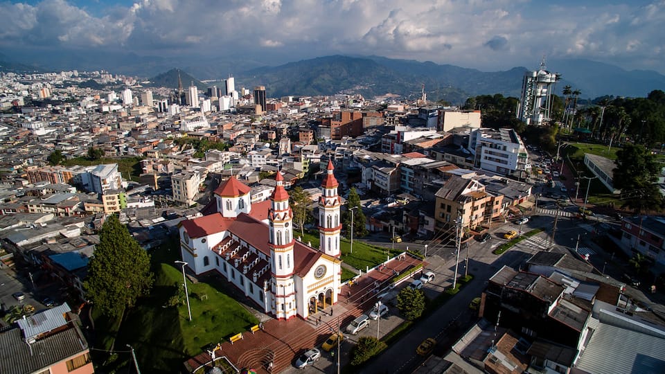 A cityscape with a prominent church featuring two towers and a large clock, surrounded by a mix of modern and older buildings, with mountains in the background.