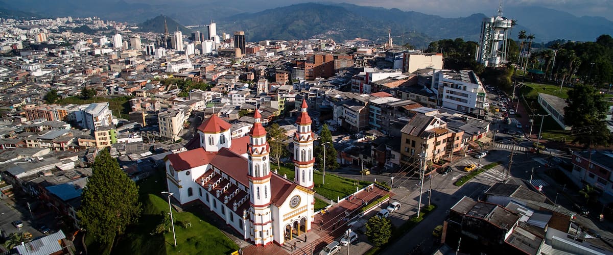 A cityscape with a prominent church featuring two towers and a large clock, surrounded by a mix of modern and older buildings, with mountains in the background.