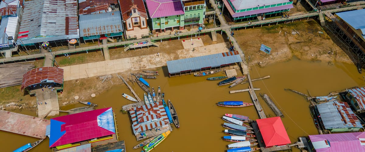 An aerial view of a riverside settlement with boats docked along the riverbanks and buildings with colorful roofs.