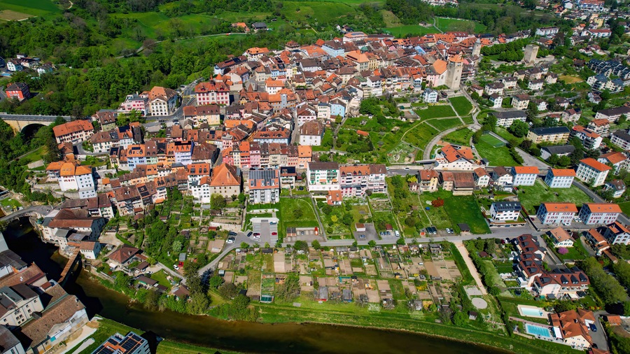 An aerial view of a town with a river, green fields, and a mix of traditional and modern buildings.