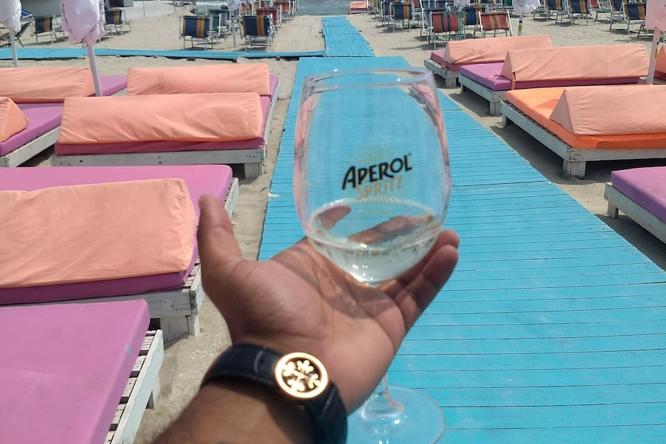A beach with rows of pink and orange lounge chairs and umbrellas, a person holding a glass of white wine, and a clear view of the ocean.