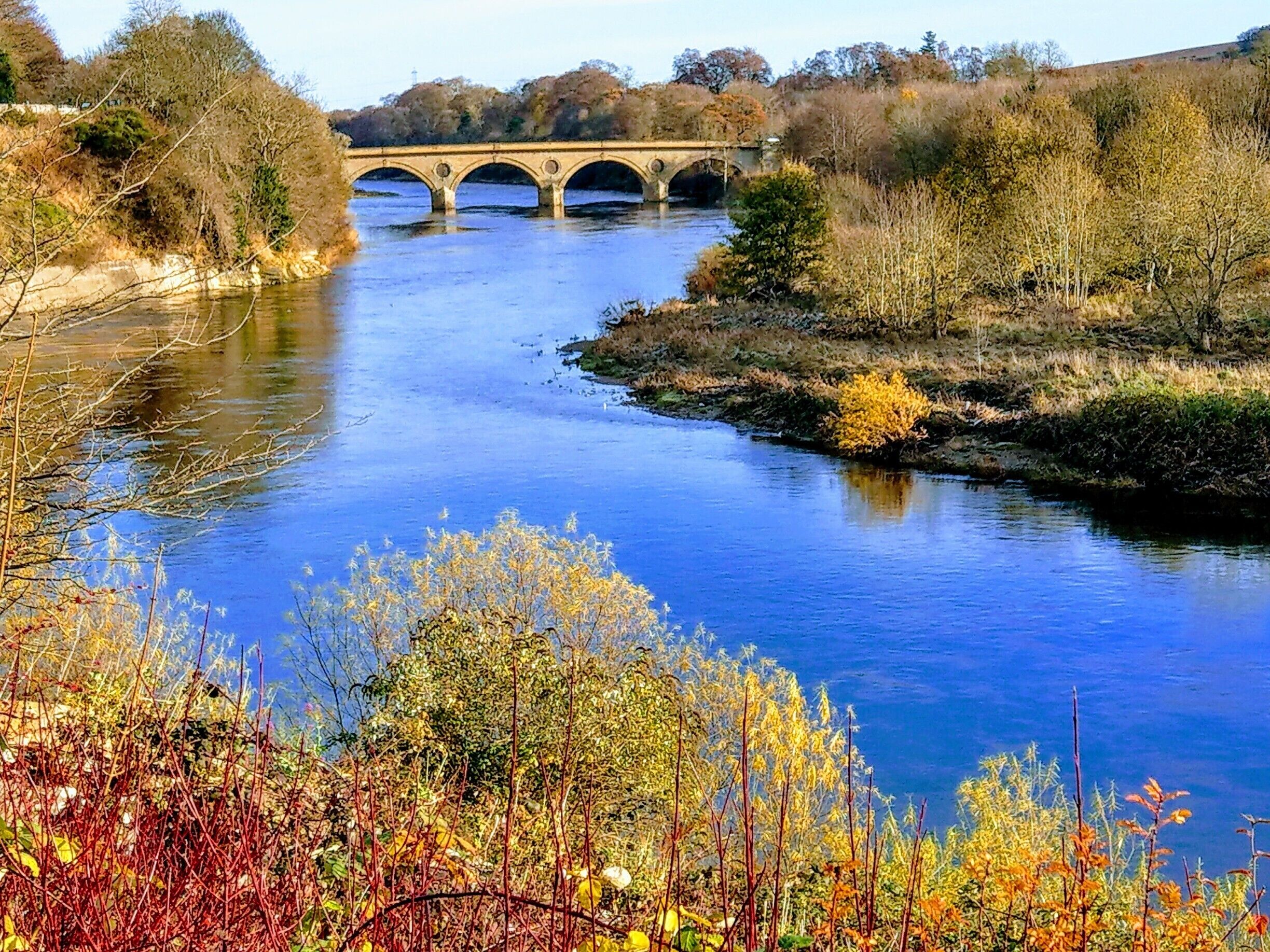 A river with a stone bridge, autumn foliage, and a hilly landscape.