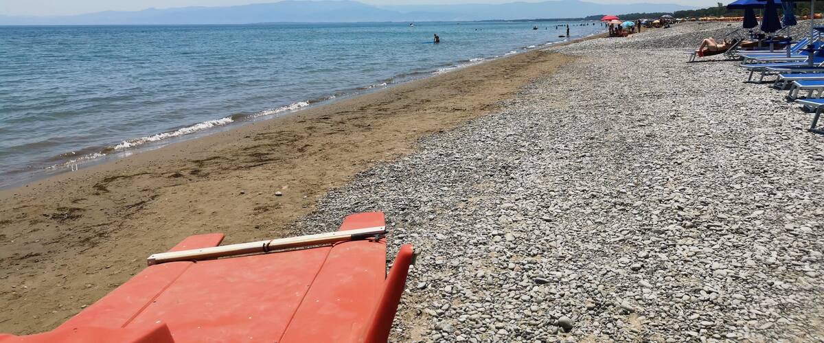 A pebbled beach with a row of orange lounge chairs and a red umbrella.