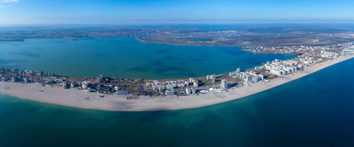 An aerial view of a coastal city with a long beachfront, a large body of water, and a distant urban area.