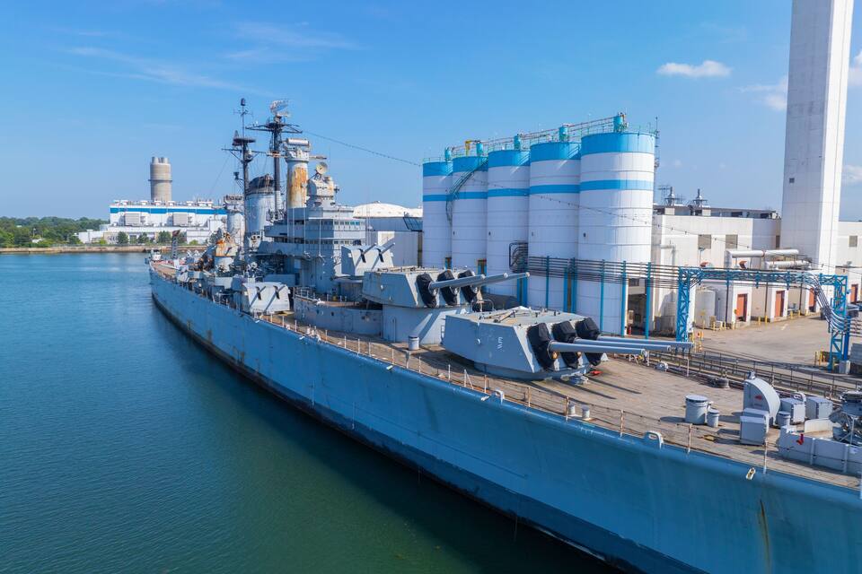 Un gran vaixell de guerra amarrat en un port, amb edificis industrials i un cel serè al fons.