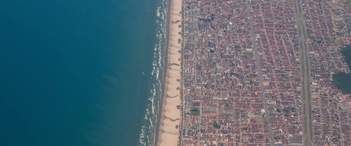 An aerial view of a coastal city with a beach, dense urban area, and a highway.