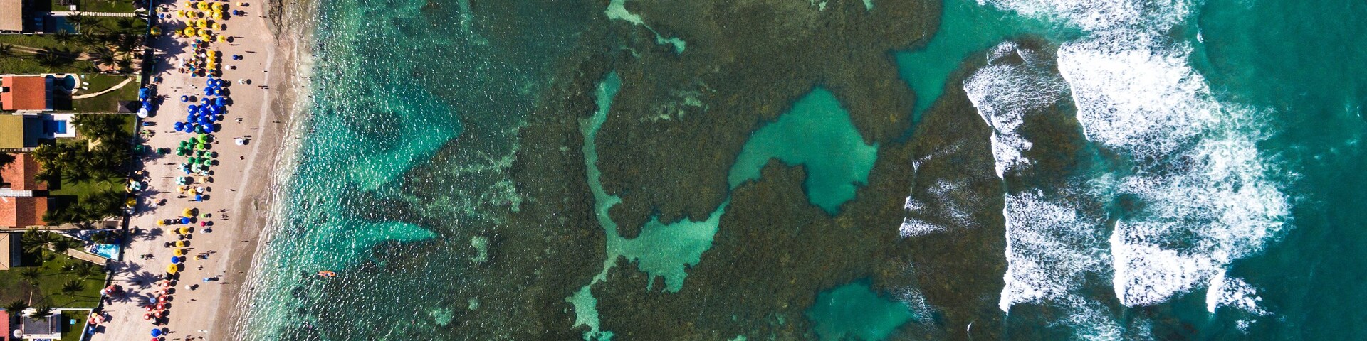 Vista aérea de uma praia com águas turquesa cristalinas, formações rochosas submersas e uma área de praia densamente povoada com guarda-sóis.